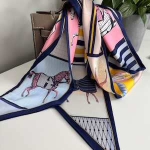 May include: A silk scarf with a navy blue border and a repeating equestrian-themed design in pink, light blue, and yellow. The scarf is draped over a brown leather handbag, with a potted plant in the background.