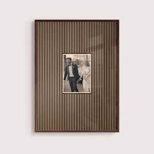 Op de afbeelding: Een rechthoekige fotolijst met een donkerbruine houten afwerking en een passe-partout met verticale strepen. In het midden bevindt zich een zwart-witfoto van een persoon in smoking en een persoon in trouwjurk die samen buiten lopen.