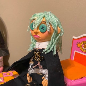 May include: A handmade doll with mint green yarn hair and teal button eyes. The doll has a peach-toned face, pink lips, and wears a black cape over a black outfit with silver metallic accents, fishnet-patterned leggings, and black boots.