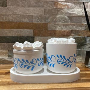 Può includere: Un set in ceramica bianca con un piccolo contenitore con coperchio a forma di fiore e un contenitore cilindrico, entrambi decorati con motivi floreali blu. Il set è posto su un vassoio bianco, ideale per il bagno.