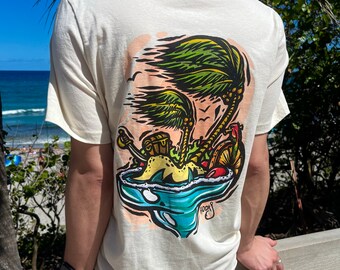 May include: A white t-shirt with a colorful graphic print of a tropical island with palm trees, a treasure chest, and a shark fin. The graphic is surrounded by a blue wave.
