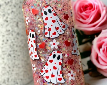 May include: A pink glitter tumbler with a green lid featuring a white ghost with red hearts pattern. The tumbler is decorated with red hearts, white flowers, and glitter.