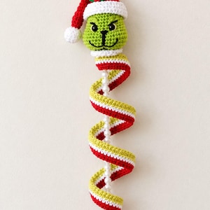 Puede incluir: Un adorno colgante de ganchillo con una cabeza de personaje verde que lleva un gorro de Papá Noel rojo y blanco con acebo. Debajo de la cabeza hay una larga cola en espiral con rayas rojas, blancas y verde lima, cuentas blancas y un cascabel.