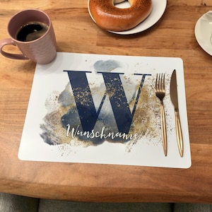 Könnte beinhalten: Ein weißer Platzteller mit einem blauen und goldenen Aquarell-Design mit dem Buchstaben "W" und dem Text "Wunschname". Eine goldene Gabel und ein Messer liegen auf dem Platzteller.