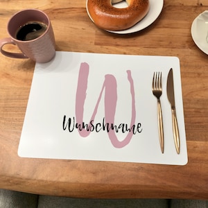 Könnte beinhalten: Ein weißer Platzdeckchen mit einem rosafarbenen 'W' und dem Text 'Wunschmaue' in Schwarz. Der Platzdeckchen liegt auf einem Holztisch mit einer Gabel und einem Messer.