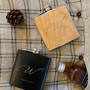 Könnte beinhalten: Zwei personalisierte Flachmänner, einer schwarz und einer aus Holz, mit einem geschriebenen Buchstaben "W" und dem Text "Wunschname" auf jedem. Die Flachmänner liegen auf einem karierten Stoffhintergrund.