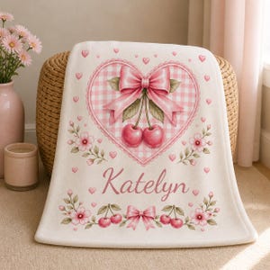 May include: A cream-colored fleece blanket featuring a central pink gingham heart with a large pink bow and two cherries. The design includes scattered small pink hearts, pink flowers, and the name Katelyn in script, with a bottom cherry border.