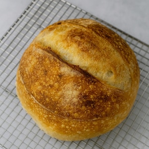 May include: A round loaf of freshly baked bread with a golden-brown crust, resting on a silver wire cooling rack. The bread has a textured surface and a diagonal score across the top, indicating it's a sourdough loaf.