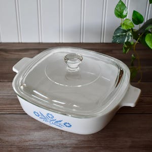 May include: White square casserole dish with a clear glass lid. The dish has a blue floral pattern on the side and two handles. The lid has a clear knob. The dish is on a wooden surface.