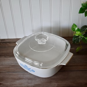 May include: White square casserole dish with a clear glass lid. The dish has a blue floral pattern on the side and two handles. The lid has a clear knob. The dish is on a wooden surface.