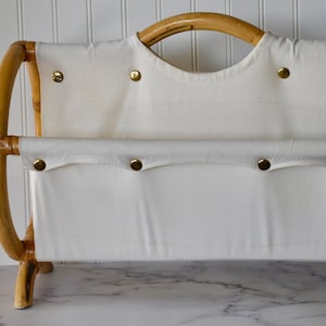 May include: A white canvas magazine rack with a bamboo frame and gold buttons. The rack has a handle on the top and stands on four legs.