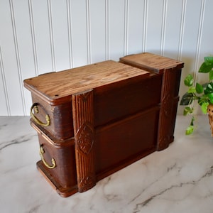 May include: A vintage wooden jewelry box with two drawers and a lift-up top. The box is made of dark brown wood with brass handles and decorative carvings. The jewelry box is sitting on a marble surface.