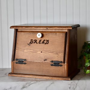 May include: A wooden bread box with a hinged front door and a white ceramic knob. The word "BREAD" is carved into the front. The box has a rustic, farmhouse style and is made of light brown wood.
