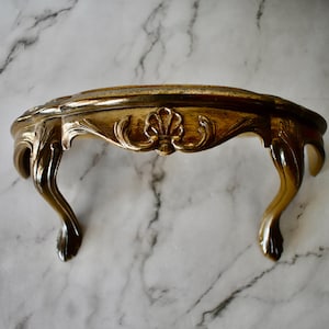 May include: Ornate, gold-colored wall shelf with a curved design. The shelf features decorative carvings and curved legs. The shelf is made of a metallic material and is mounted on a marble-patterned wall. A decorative piece for home decor.