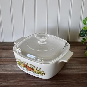 May include: A white casserole dish with a clear glass lid. The dish has a square shape with rounded corners and side handles. It features a colorful botanical design with the text "Le Persil, La Sauge".
