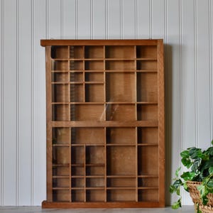 May include: A rectangular wooden display case featuring a glass front and numerous small, varied compartments arranged in a grid pattern. The frame is constructed from medium-toned brown wood, providing a structured storage solution for items.