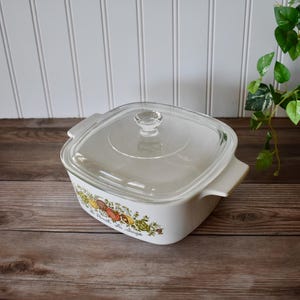 May include: A square white ceramic casserole dish with integrated side handles and a clear glass lid. The exterior features a colorful botanical illustration of vegetables and herbs with the text La Persillade Au Sauge printed on the front.