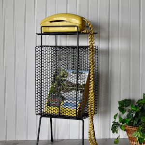 May include: A black metal telephone stand with a perforated mesh body and a top shelf. A yellow landline telephone with a long coiled cord sits on the top shelf, and a magazine is stored inside the mesh compartment below.