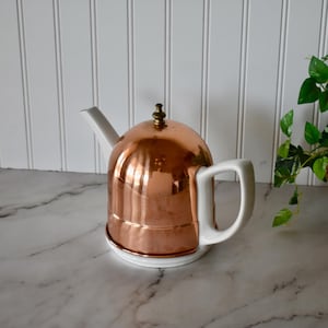 May include: A teapot featuring a white ceramic base, handle, and spout, combined with a polished copper dome-shaped cover. The copper lid is topped with a small, metallic finial, creating a contrast between the white ceramic and the metallic copper.