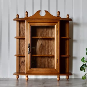 May include: A medium-toned wooden wall cabinet featuring a central glass-paneled door with a decorative metal pull handle. The unit includes three open shelves on each side and a curved top crest with small finials, supported by four ball feet.