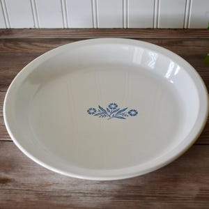 May include: A round white ceramic pie dish with a smooth, glossy finish. The center of the dish features a small, detailed blue floral pattern consisting of two stylized flowers and leaves arranged in a symmetrical, horizontal cluster design.