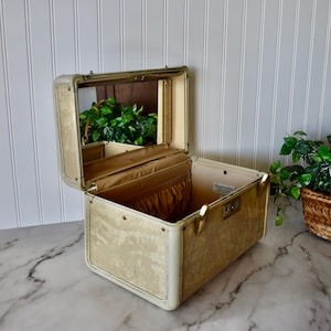May include: Open vintage vanity case with a gold and cream floral pattern. The case has a mirror inside the lid and gold-toned hardware. The interior features a small pocket and a pleated fabric lining.