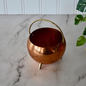 May include: A polished copper-colored metal bowl with a spherical body, featuring a thin, arched gold-toned metal handle attached to the rim and three small, pointed gold-toned metal feet at the base, creating a tripod-style support structure.