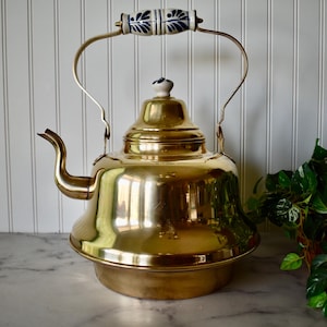May include: A shiny brass kettle with a blue and white ceramic handle and knob. The kettle has a rounded body and a spout on the side.