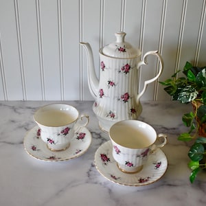 May include: A white tea set with a floral pattern. The set includes a teapot, two cups, and two saucers. The teapot has a lid and a handle. The cups and saucers have a floral pattern with pink flowers. The set is on a white marble surface.