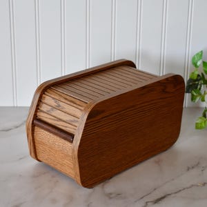 May include: A wooden bread box with a roll-top lid. The box is made of a light brown wood with a darker brown handle. The lid has a series of horizontal slats. The bread box is on a marble surface.