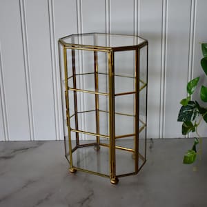 May include: A hexagonal display case featuring clear glass panels framed in a metallic gold finish. The structure includes a hinged door with a small latch, multiple interior glass shelves, and is supported by four small, rounded gold feet.