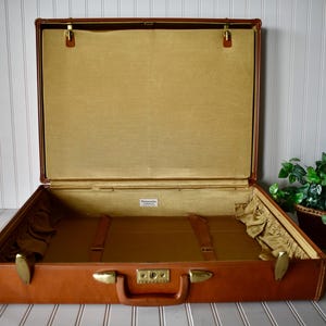 May include: Open vintage brown suitcase with brass hardware. The interior is lined with a gold fabric and includes two brown straps. The suitcase has a handle and a lock. The brand name is visible on a small tag inside.