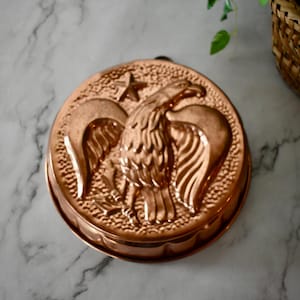 May include: A round copper mold featuring a raised relief design of an eagle with spread wings and a five-pointed star above its head. The background of the mold has a textured, pebbled surface, and the edges are scalloped.