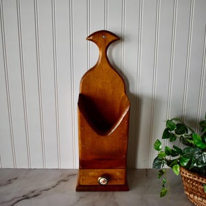 May include: A tall, wall-mounted wooden storage bin with a medium brown finish. The unit features a curved top with a small hole for hanging, an open-front compartment for storage, and a single small drawer at the base with a round floral knob.