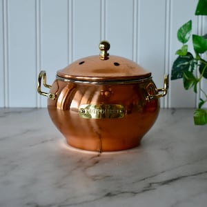 May include: A copper potpourri pot with a lid and brass handles. The pot has a round shape and a shiny finish. The lid has small holes and a brass knob. The pot has a brass label that reads "POTPOURRI".