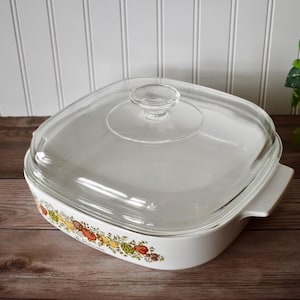 May include: White square casserole dish with a clear glass lid. The dish has a decorative floral pattern around the rim. The lid has a small, round handle. The dish is on a wooden surface.