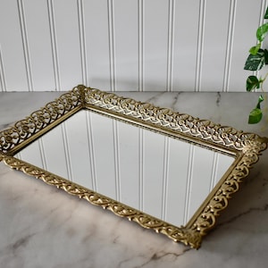 May include: A rectangular gold-toned vanity tray with a mirrored surface. The tray has an ornate, filigree-style border. The tray is resting on a marble surface, with a white paneled wall in the background.