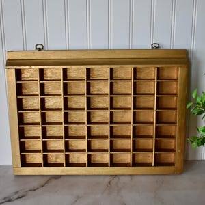 May include: A rectangular gold-painted wooden display case featuring a grid of forty-eight small, uniform square compartments. The unit includes a flat top ledge and two metal hanging loops attached to the upper frame for wall mounting purposes.