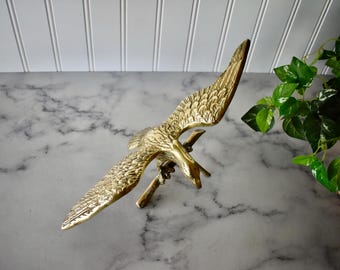 May include: A golden brass eagle sculpture with outstretched wings, perched on a branch. The bird's feathers are intricately detailed. The sculpture is set against a marble surface with a green plant in the background.