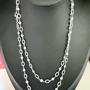 May include: A silver necklace with two strands, displayed on a black mannequin. The necklace features a repeating pattern of oval and round clear stones. The necklace is a jewelry item.