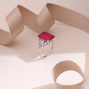 May include: A silver ring with a large, rectangular, ruby-red gemstone. The ring features a detailed, Art Deco-inspired design with smaller, clear gemstones. A beige ribbon is in the background, adding a touch of elegance to the jewelry.