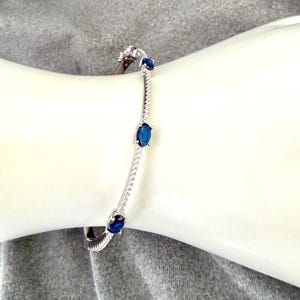 May include: A silver bracelet with three oval, dark blue gemstones. The bracelet is adorned with small, clear stones and is displayed on a white surface against a gray background.