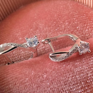 May include: A pair of silver-toned hoop earrings featuring an infinity-shaped design. Each earring is accented with a single clear round-cut stone at the top and a row of small pavé-set stones along the curved band, finished with a hinged clasp.