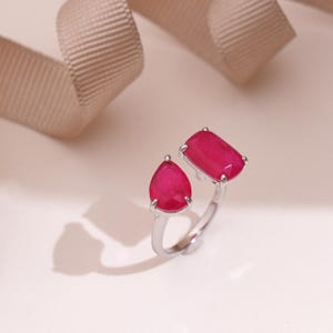 May include: A silver ring featuring two vibrant, ruby-red gemstones. One stone is a pear shape, the other a rectangular shape. The ring's design is open, with the gemstones set on either side of the band. The ring is displayed on a light-colored surface.