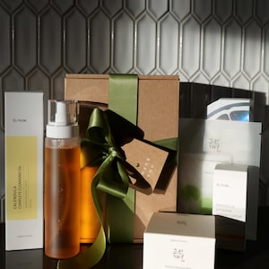 Puede incluir: Una colecci&oacute;n de productos para el cuidado de la piel, incluyendo aceite limpiador, s&eacute;rum y mascarillas faciales, dispuestos sobre una superficie oscura. Los productos est&aacute;n empaquetados en cajas blancas y amarillas, con una caja de regalo atada con una cinta verde.