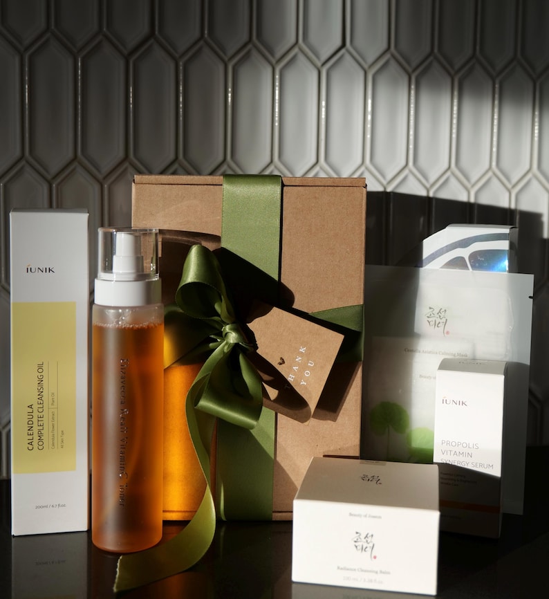Puede incluir: Una colecci&oacute;n de productos para el cuidado de la piel, incluyendo aceite limpiador, s&eacute;rum y mascarillas faciales, dispuestos sobre una superficie oscura. Los productos est&aacute;n empaquetados en cajas blancas y amarillas, con una caja de regalo atada con una cinta verde.