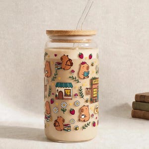 May include: A clear glass tumbler with a bamboo lid and a glass straw, filled with a beige liquid. The exterior features colourful illustrations of capybaras, small buildings, flowers, strawberries, bees, and books scattered across the surface.
