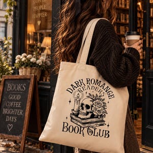 Peut inclure: Un sac fourre-tout en toile beige avec une impression graphique noire représentant un crâne humain, une bougie allumée et des roses posées sur une pile de livres. Le texte indique Dark Romance Readers Book Club. Le sac a deux longues anses.