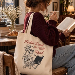 Peut inclure: Un sac fourre-tout en toile couleur crème avec une impression graphique noire et rouge représentant un squelette portant une couronne de fleurs et lisant un livre. Le texte Feelin' Dead But Well Read est écrit en rouge.