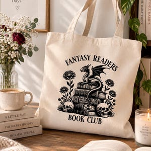 Peut inclure: Un sac fourre-tout en toile couleur crème avec une impression graphique noire représentant un dragon perché sur une pile de livres, entouré de fleurs et de deux crânes. Le texte "FANTASY READERS BOOK CLUB" est imprimé en arc au-dessus et en dessous.
